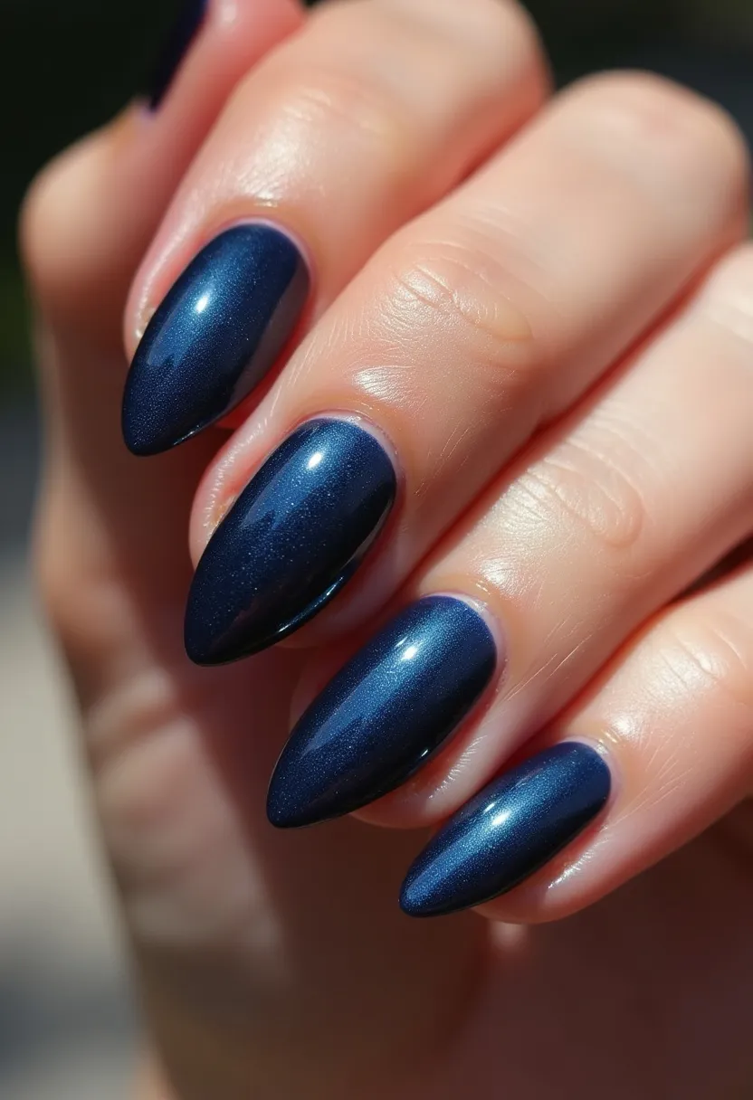 The nail design showcases a sophisticated and elegant look featuring a dark, metallic blue color palette. The nails are shaped into a sharp almond form, elongated and tapering to a precise point. The polished finish suggests a high glossy effect typically achieved with gel or shellac nail treatments, providing both durability and a lustrous shine. The color is uniform across all nails, creating a cohesive and refined appearance without additional intricate patterns or decorations. This design, with its deep blue hue, is versatile but particularly fitting for the winter season or formal events, exuding a classic and polished aesthetic.